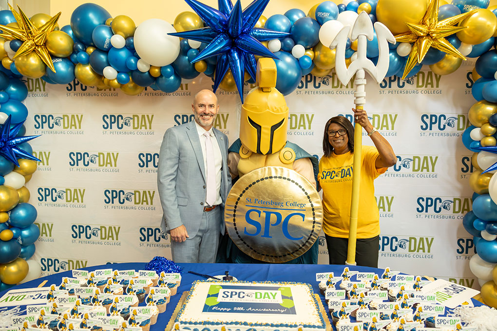 Trustee Jason Butts, Titus the Titan, and Dr. Tonjua Williams, at SPC Day 2025, posing in front of blue and gold balloons and behind the SPC Day cake and cupcakes.