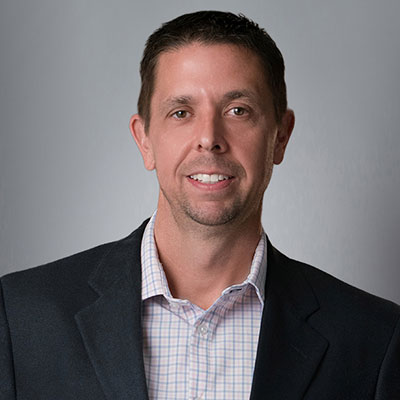 A Portrait of Richard Franz III, wearing a black suit coat over a white dress shirt with blue vertical and horizontal stripes.