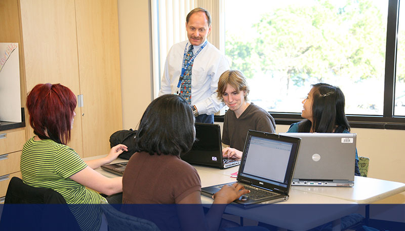 male professor in a tie teaching students sitting at a table in front of laptop computers