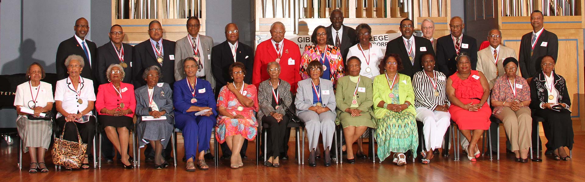 A group photo of the Gibbs Junior College Alumni Association members, one row seated and one row standing, on a stage, in front of a pipe organ.