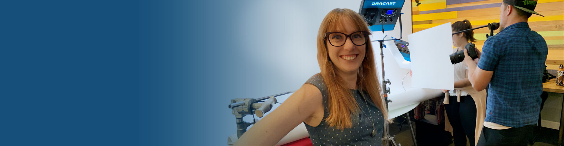 Blue gradient image that blends into a female student smiling at the camera while two students work in the background on digital media equipment.