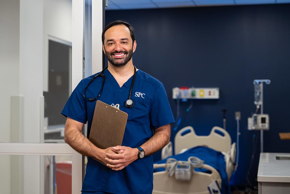 male student wearing blue scrubs holding a clipboard with his hands folded
