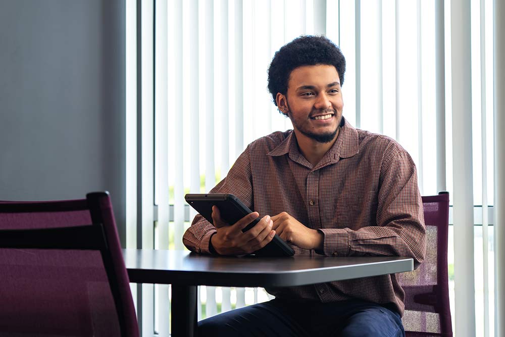 male student in a brown button down shirt holding a tablet and smiling wide