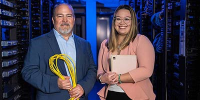 photo of male professor in a blue blazer and female student in pink jacket standing with computer cables standing in front of a room of servers